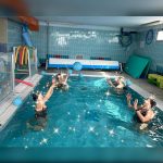 Persone impegnate in esercizi di gruppo in piscina durante una seduta di idrochinesiterapia con palloni galleggianti.