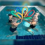 Gruppo di persone durante una lezione di acquagym in piscina con tubi galleggianti colorati utilizzati per esercizi di coordinazione e benessere fisico.