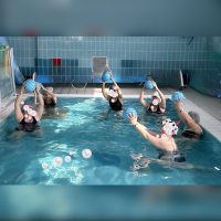 Gruppo di persone durante esercizi di idrochinesiterapia in piscina terapeutica con palloni galleggianti.