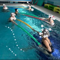 Gruppo di persone durante esercizi di idrochinesiterapia in piscina con noodles galleggianti colorati.